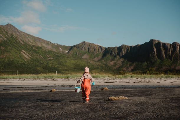 Bilde av jente fra familiefoto som går med en bøtte på en strand, med høye fjell i bakgrunnen