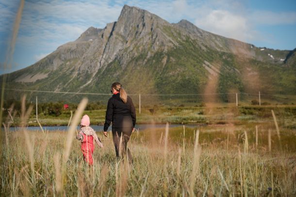 Familiefoto av mor og datter på en eng med strå i forgrunnen og høye fjell i bakgrunnen